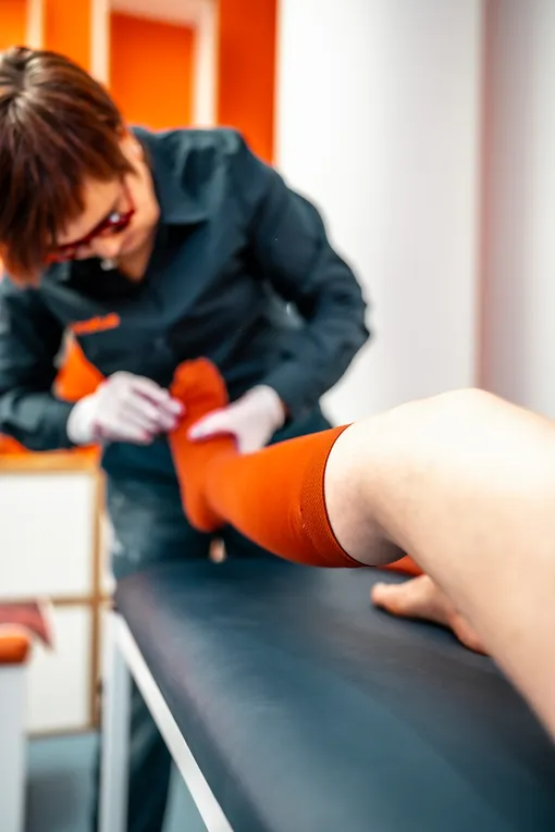 Die Beraterin zieht mit Hilfe von Handschuhen die orangefarbenen Kompressionsstrümpfe an der Kundin an